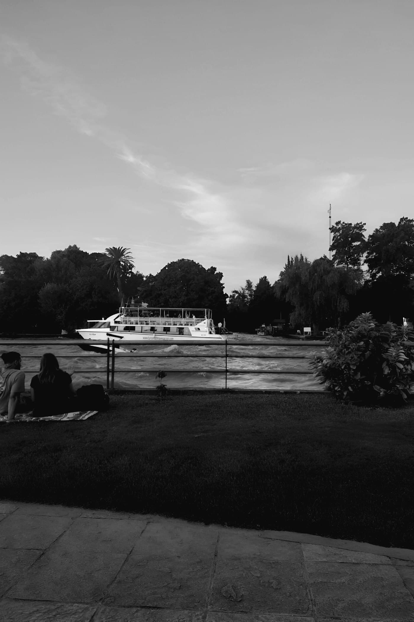 A river at dusk, a boat passing, people sitting on the grass.